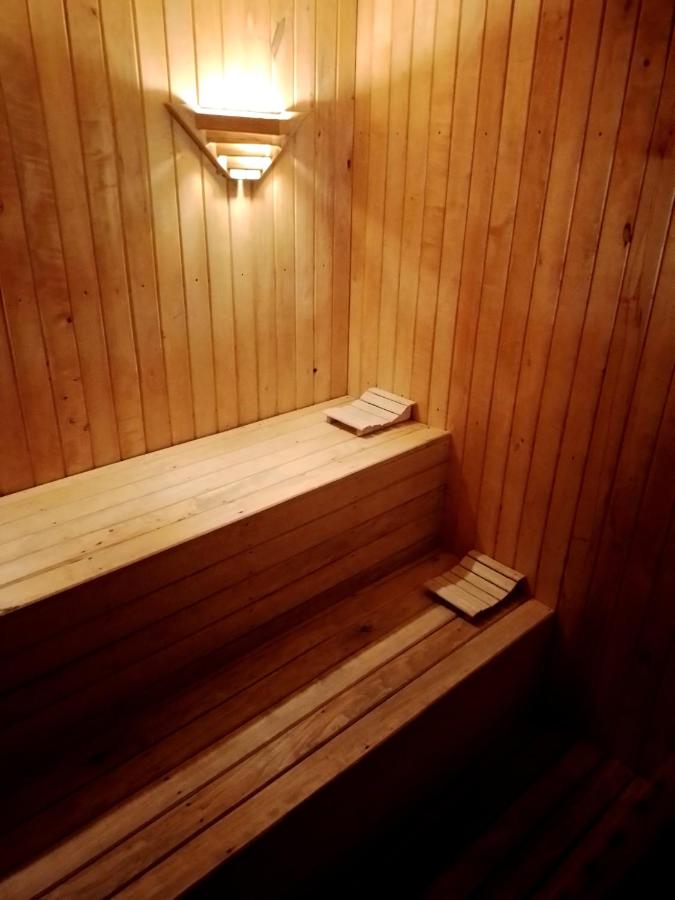 A wooden sauna interior is showcased, featuring smooth, light-colored wood paneling. Two tiered benches made of slatted wood are visible, providing ample seating. Soft, warm lighting from a wall-mounted fixture creates a calming ambiance.