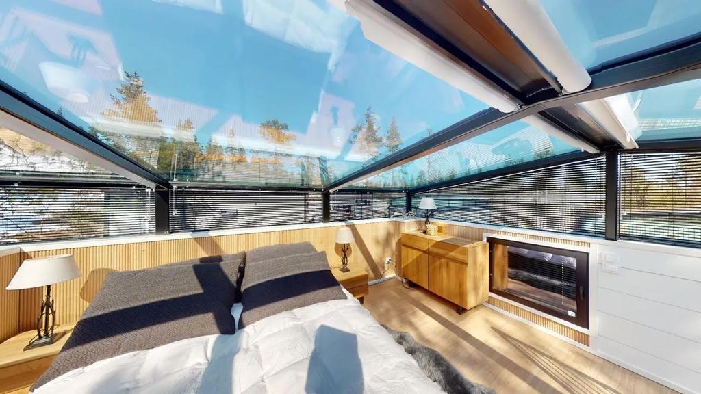 A unique space features a glass ceiling allowing natural light to flood the room. Two spacious beds are positioned side by side, complemented by wooden nightstands with lamps. A modern console is visible, and a view of trees can be seen through the expansive windows.