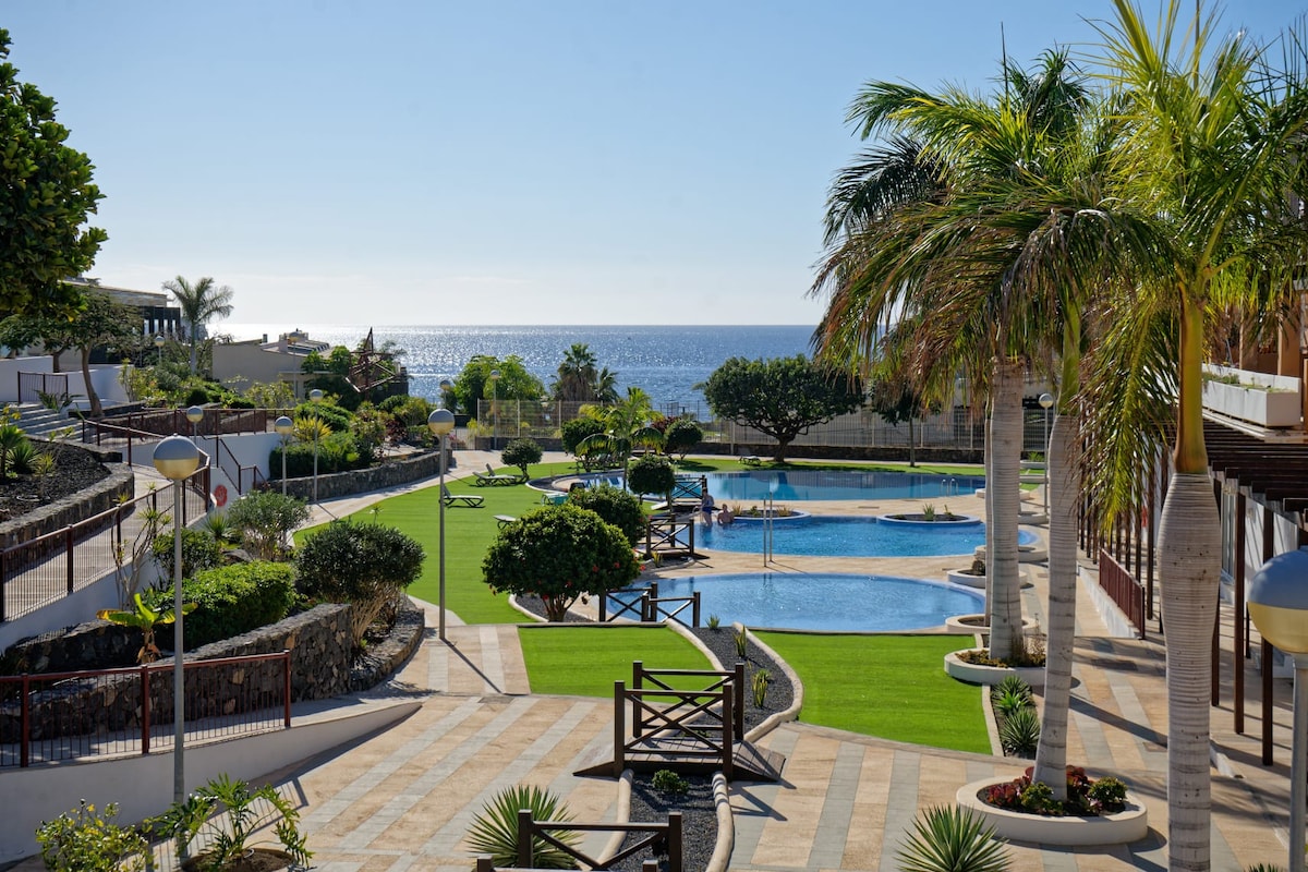 The image presents a vibrant landscape featuring multiple swimming pools surrounded by lush gardens and palm trees. The serene sea can be seen in the background, creating a tranquil atmosphere. Well-maintained pathways and green lawns invite leisurely strolls through the complex.
