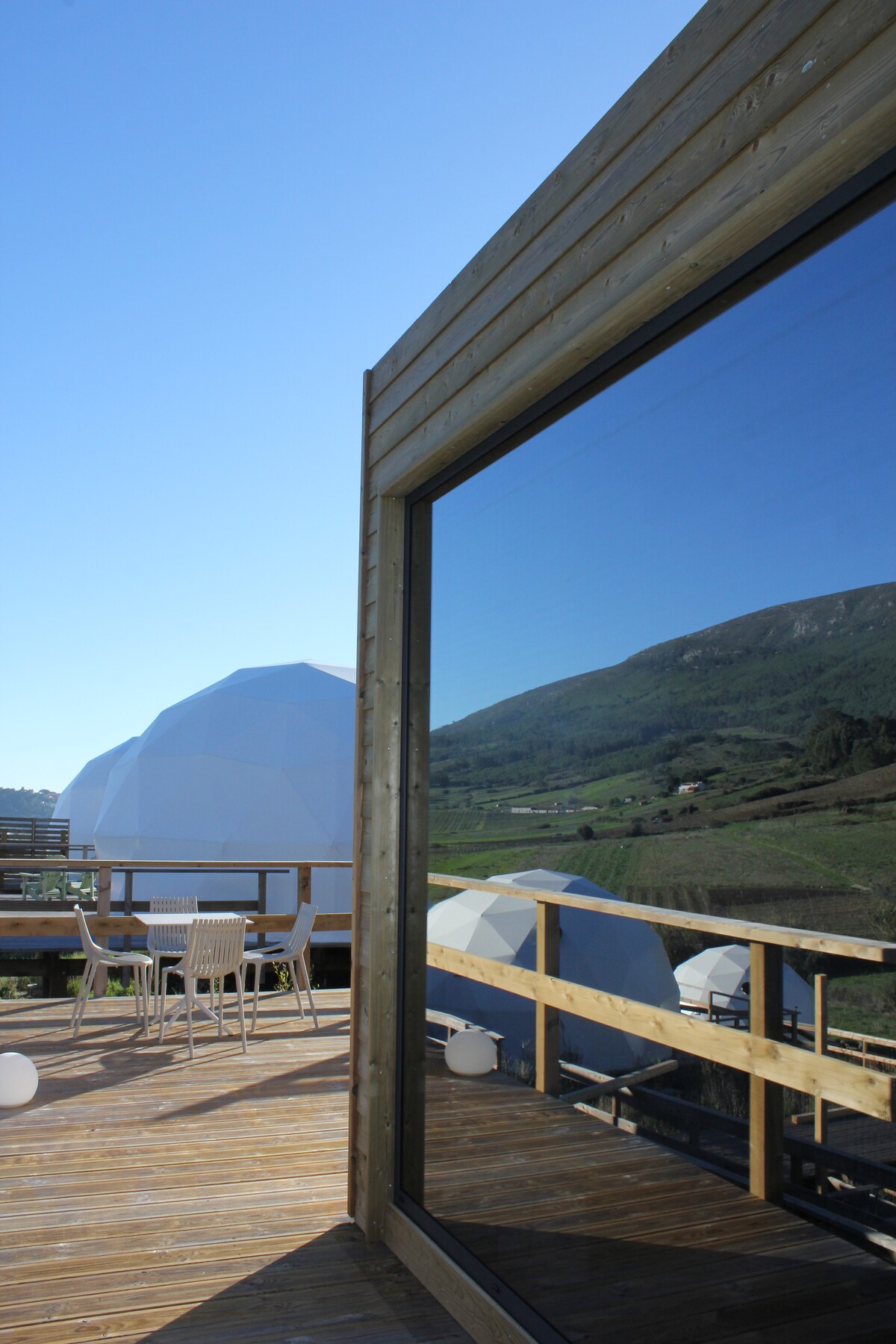 A wooden terrace features a panoramic view of the surrounding hills, complemented by a reflective glass wall of the bungalow. Modern outdoor furniture is arranged on the deck, with geometric domes visible in the background under a clear blue sky.
