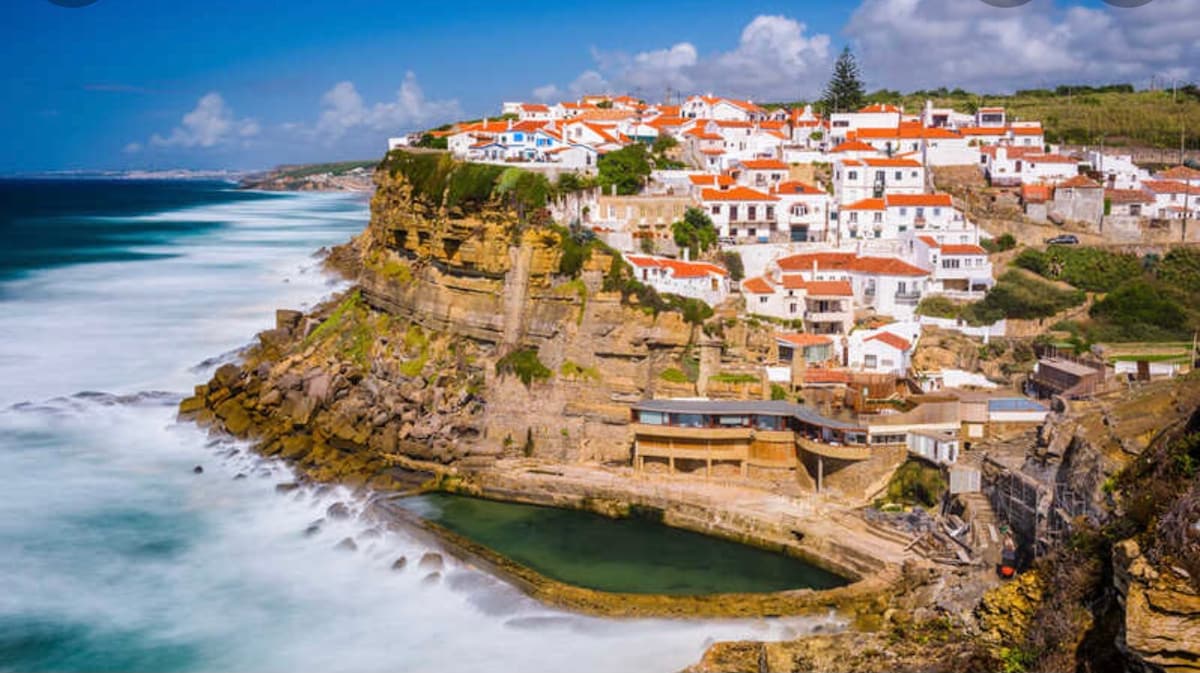 The image captures a coastal village perched on a cliff, showcasing buildings with red roofs against a backdrop of blue skies. Waves gently crash against the rocky shore, and a secluded pool is visible at the base of the cliff, framed by the surrounding landscape.