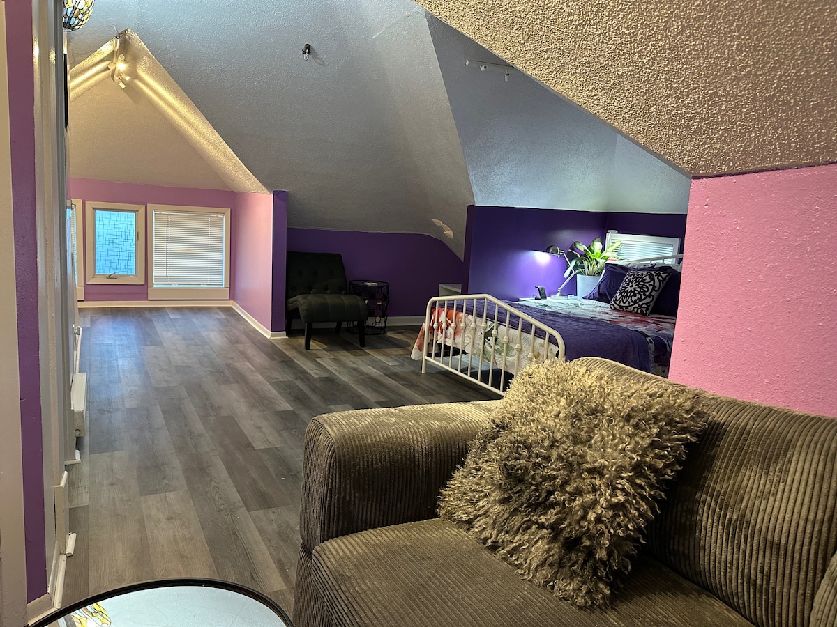 The open attic space features two queen beds adorned with decorative pillows and vibrant wall art. A cozy seating area with a sofa enhances the room's layout. Natural light enters through the windows, illuminating the colorful walls and wooden flooring.