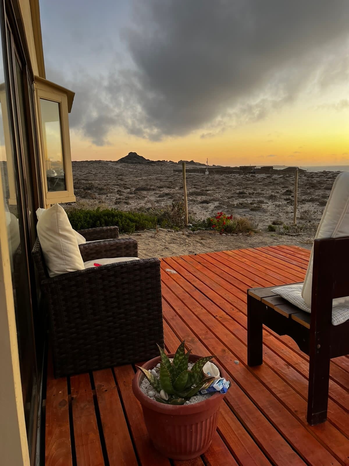 A seating area is seen on a wooden deck, featuring a wicker chair with a white cushion and a potted plant. In the background, rocky terrain leads to a horizon where the sun is setting, casting a warm glow over the landscape.