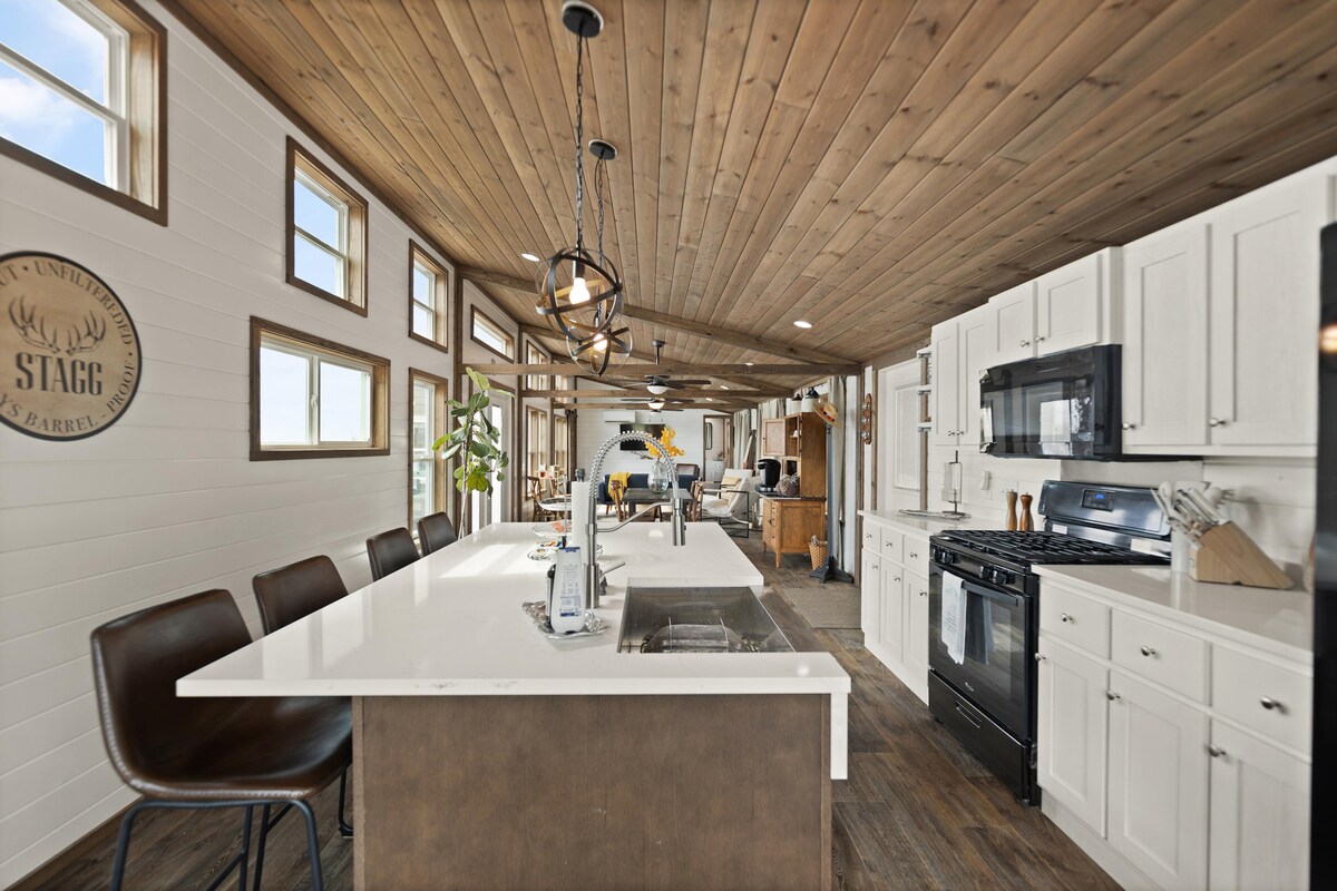 An open kitchen featuring a large island with seating, surrounded by modern cabinetry and white countertops. Stainless steel appliances, including a gas stove and microwave, are visible. Natural light fills the space through multiple windows, highlighting the wood beam ceiling and creating a welcoming environment.