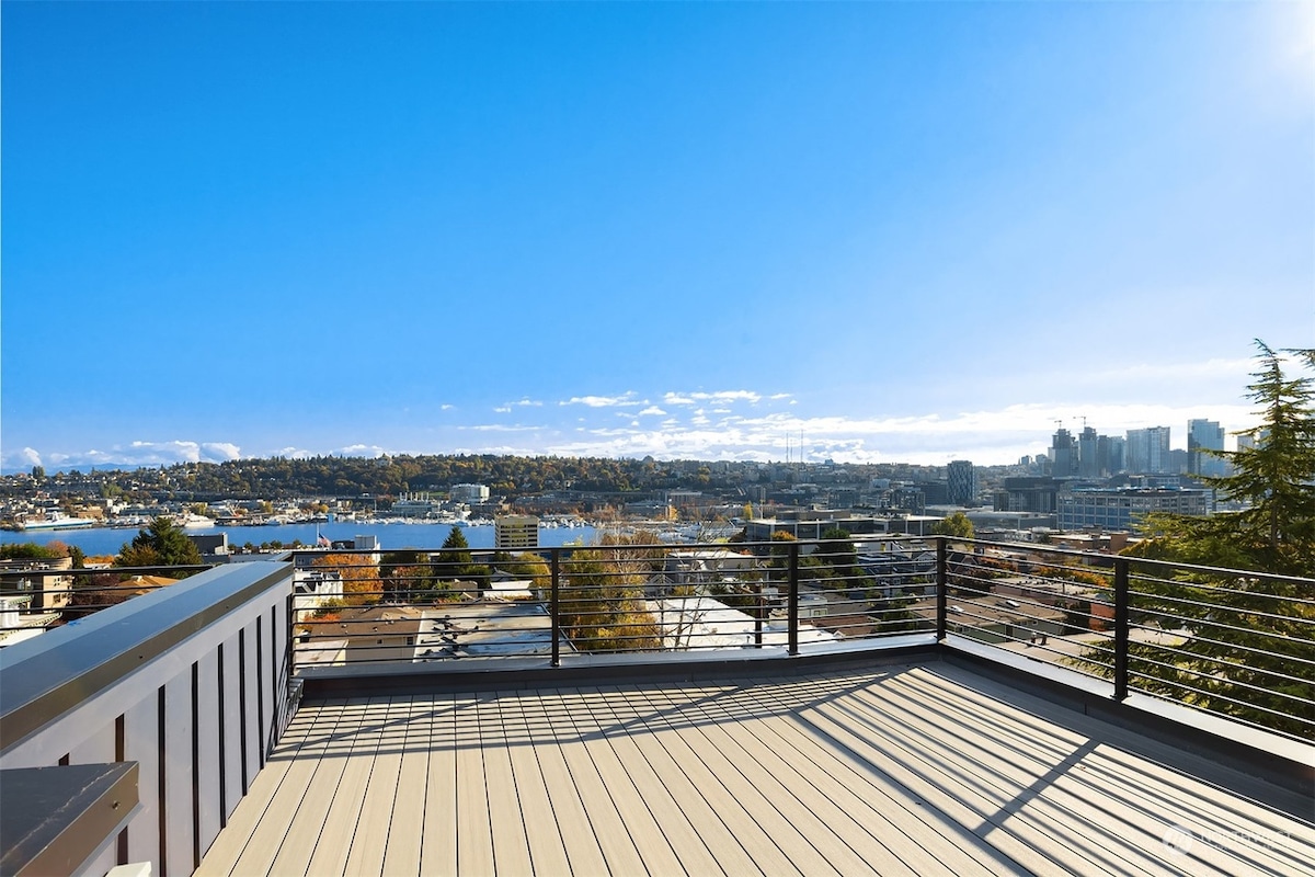 A spacious rooftop terrace offers panoramic views of Lake Union and the Seattle skyline under a clear blue sky. The deck features wooden flooring and a railing providing safety while enjoying the open outdoor space.