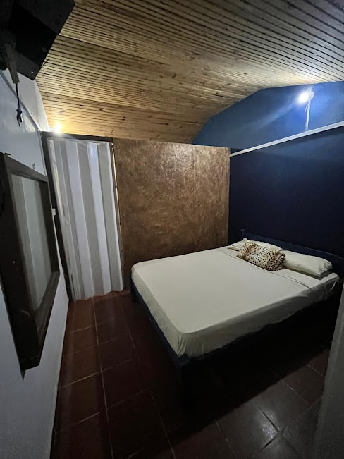 A compact sleeping area is presented with a spacious bed adorned with a decorative pillow. The room features a wooden ceiling and a dark blue wall. A framed mirror is positioned near the bed, reflecting the room's subtle lighting.