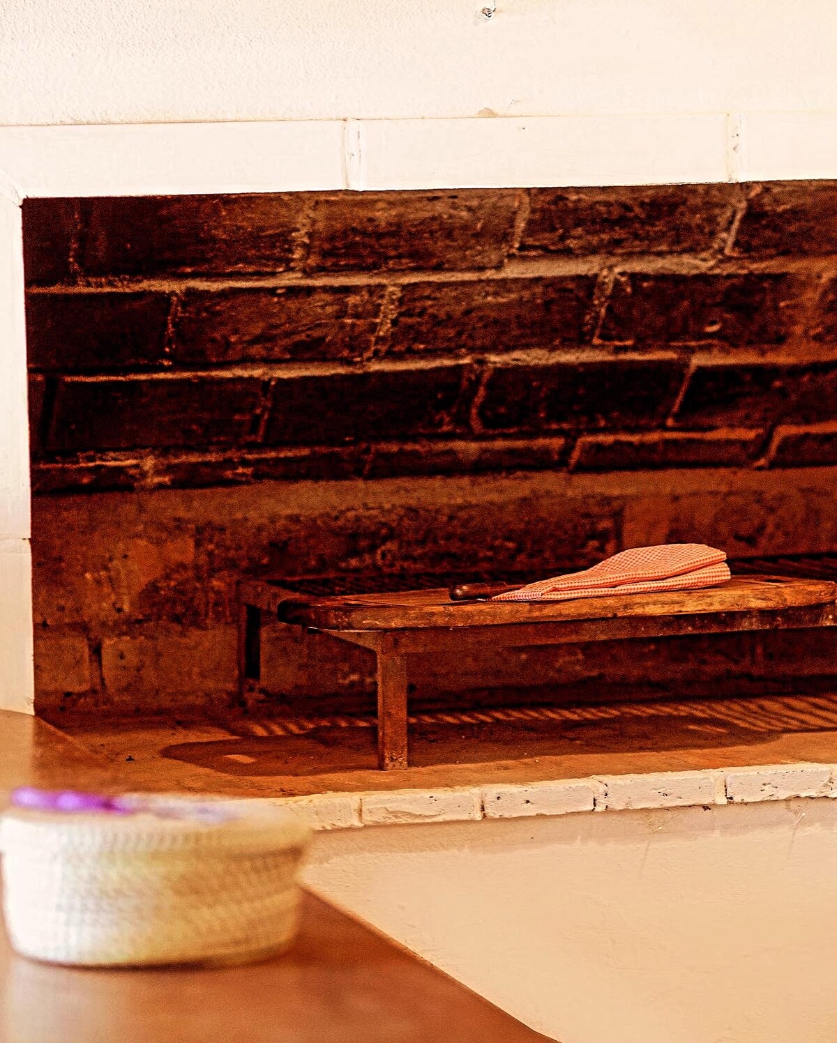 An internal BBQ grill is positioned against a rustic brick wall, showcasing a simple metal frame with a wooden surface. A partially folded apron is placed on top, adding a practical element. The surrounding area features a smooth, neutral-toned countertop.