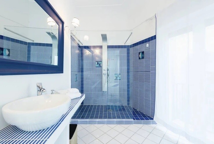 A spacious bathroom is highlighted by a walk-in shower with blue tiled walls and a patterned floor. A modern sink is positioned on a minimalist counter, complemented by a large mirror above. Natural light filters in through sheer curtains, enhancing the room's fresh ambiance.