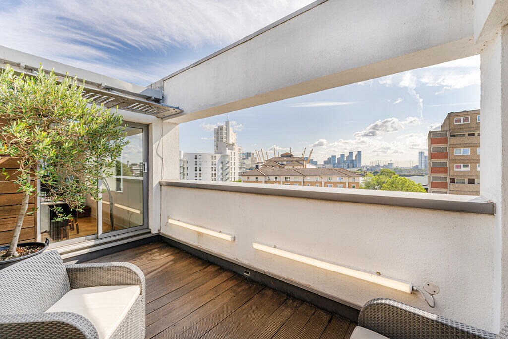 An outdoor terrace is shown, featuring a wooden deck with two woven lounge chairs. A small potted plant adds a touch of greenery. The view includes city buildings and a clear sky, creating a spacious and open atmosphere.
