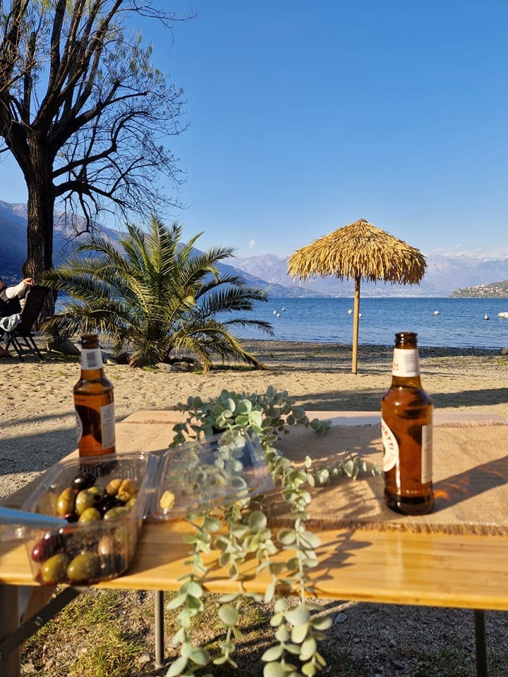 A sandy beach area is visible, featuring two bottles of beer on a wooden table. In the background, a straw umbrella provides shade. Lush palm trees and mountains frame the serene lake, creating a peaceful atmosphere. A clear blue sky complements the tranquil setting.