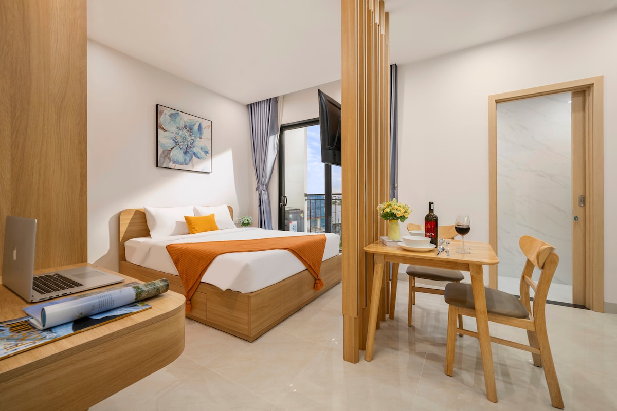 A modern apartment interior features a spacious bed dressed in crisp white linens, complemented by an orange throw. A wooden dining table with two chairs sits across from a work desk, alongside a large window allowing natural light and a view of the balcony.