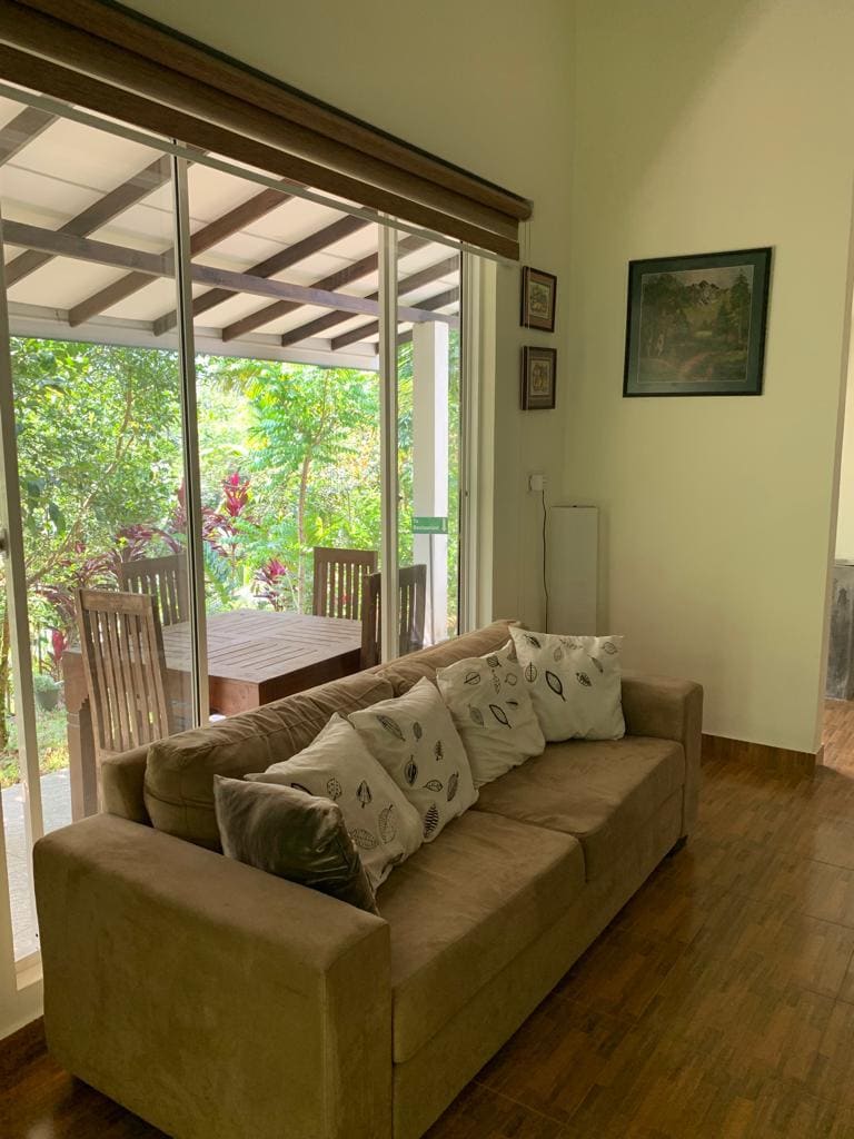 A comfortable seating area is presented with a beige sofa adorned with decorative pillows. Large glass windows allow natural light to filter in, offering views of a lush garden and a dining set located on a covered patio just outside.