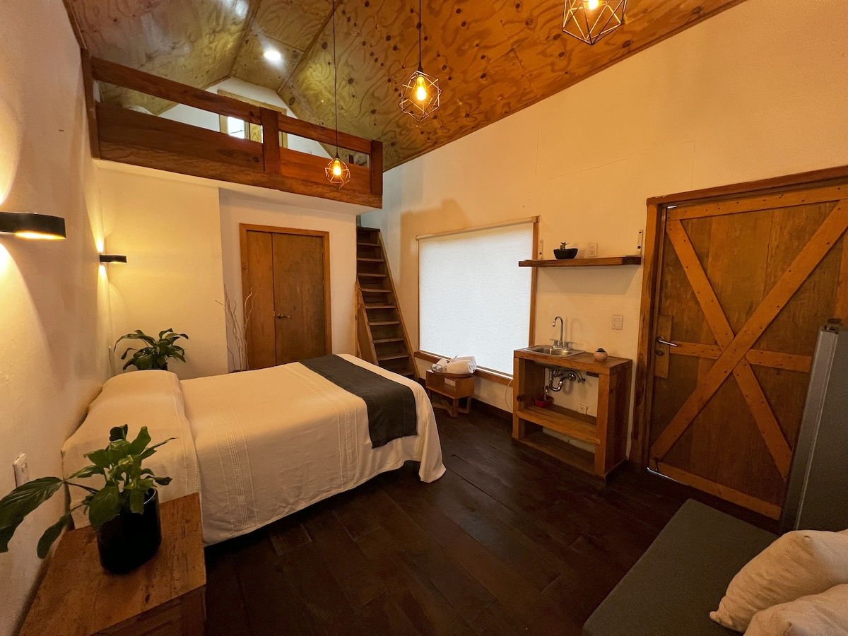 A cozy interior features a spacious bed with neutral bedding and a wooden headboard. Soft lighting is provided by hanging fixtures. A small kitchenette is present, along with a comfortable seating area. A staircase leads to a loft space, adding character to the room.