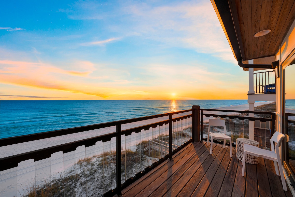 A private balcony overlooks the sandy beach and Gulf waters, with a stunning view of the sunset. White outdoor chairs are positioned on the wooden deck, providing a cozy spot to enjoy the coastal scenery.