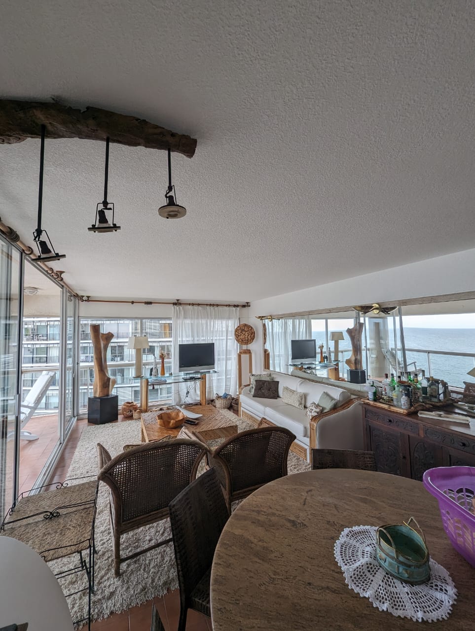 A bright living room space features large glass windows offering ocean views. A comfortable sofa is positioned alongside a wooden coffee table. Woven chairs surround a dining table. Decorative elements such as sculptures and a large mirror are visible, enhancing the room's decor.