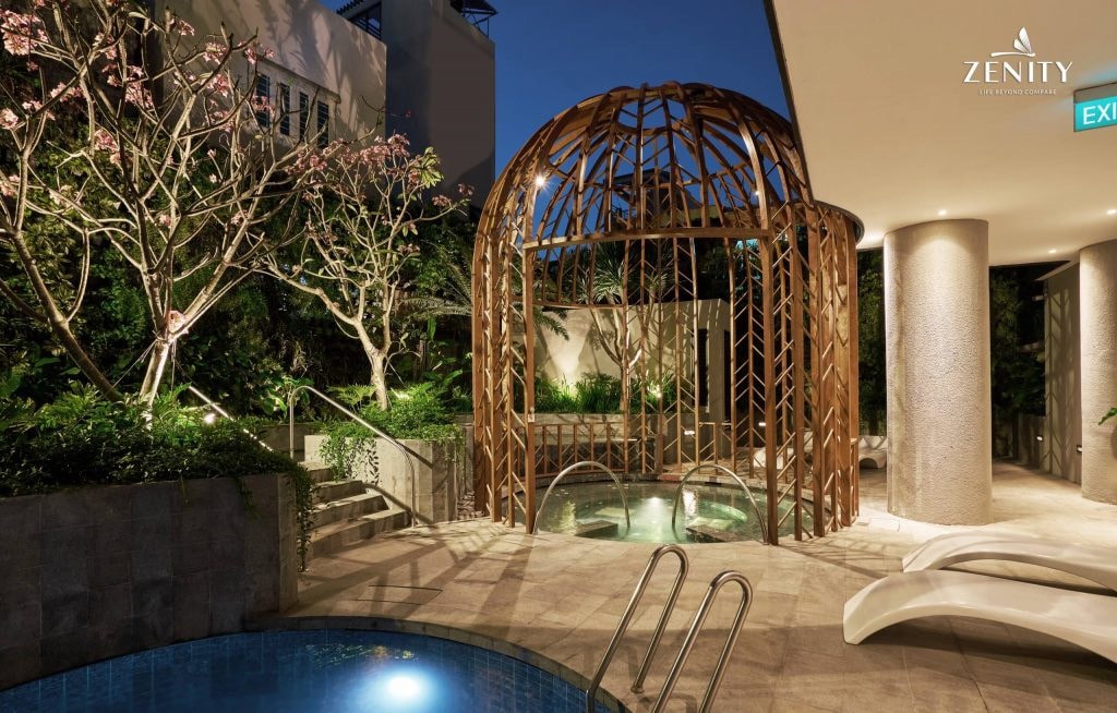 An outdoor relaxation area is illuminated by soft lighting, featuring a unique wooden structure that provides shelter. Two hot tubs are positioned nearby, surrounded by lush greenery and manicured plants, creating a serene environment for guests to unwind.