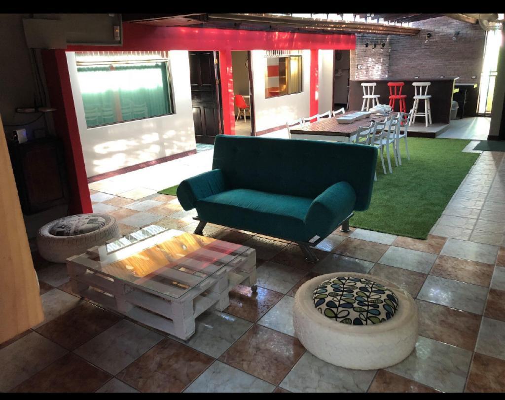 An inviting common area displays a green sofa and a coffee table made of pallets. The floor features a mix of tile and grass-like carpet, with white chairs arranged around a long table in the background. Large windows allow natural light to fill the space.
