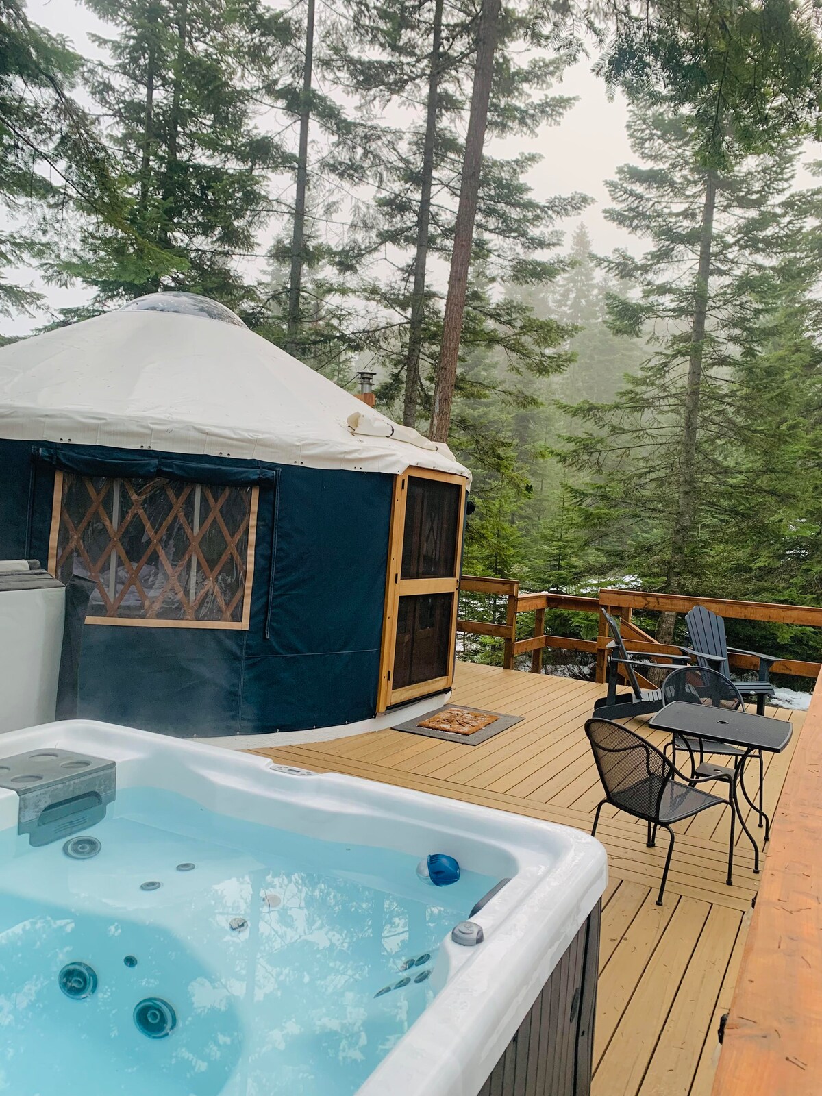 A hot tub bubbles gently on a wooden deck, surrounded by towering trees. The yurt structure is visible in the background, featuring a wooden door and lattice window. Outdoor seating options include a small round table with chairs and a lounge chair, inviting relaxation in a natural setting.