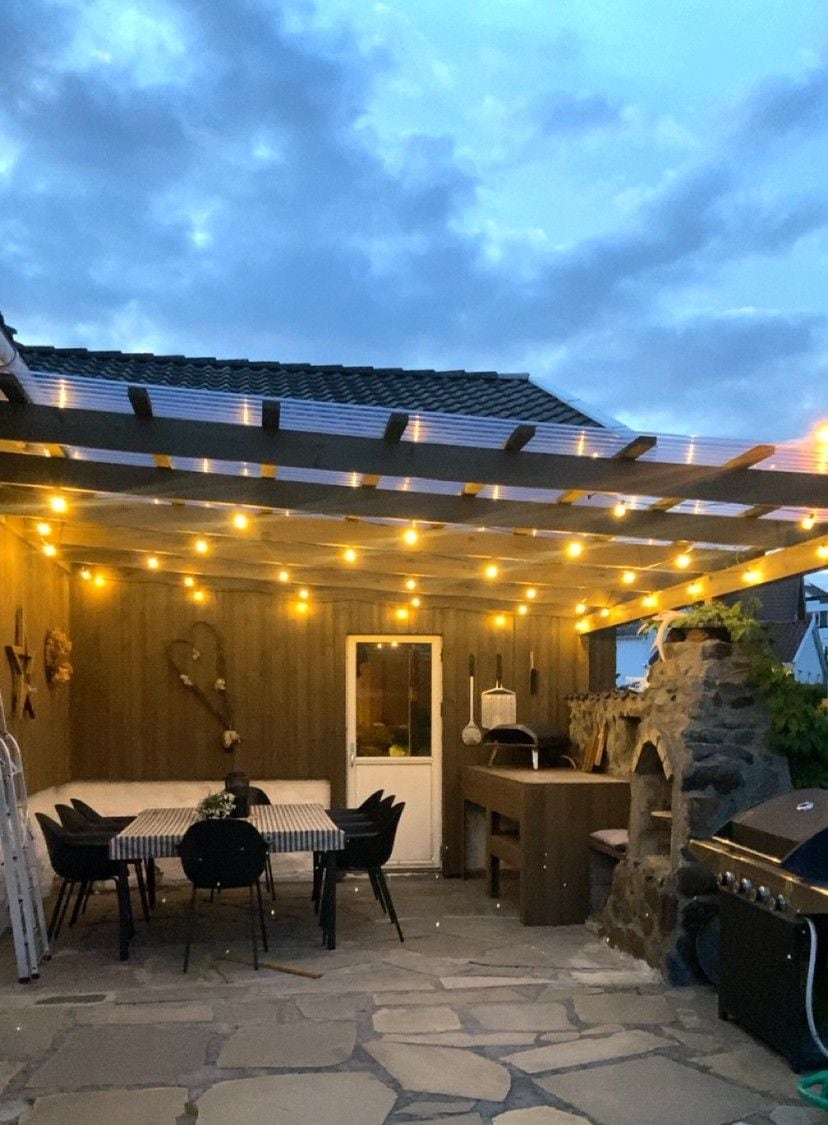 An outdoor patio area is featured, illuminated by warm string lights. A wooden table is surrounded by black chairs, and a barbecue grill is positioned in the corner. Natural stone elements enhance the space, creating a cozy atmosphere for gatherings on clear evenings.