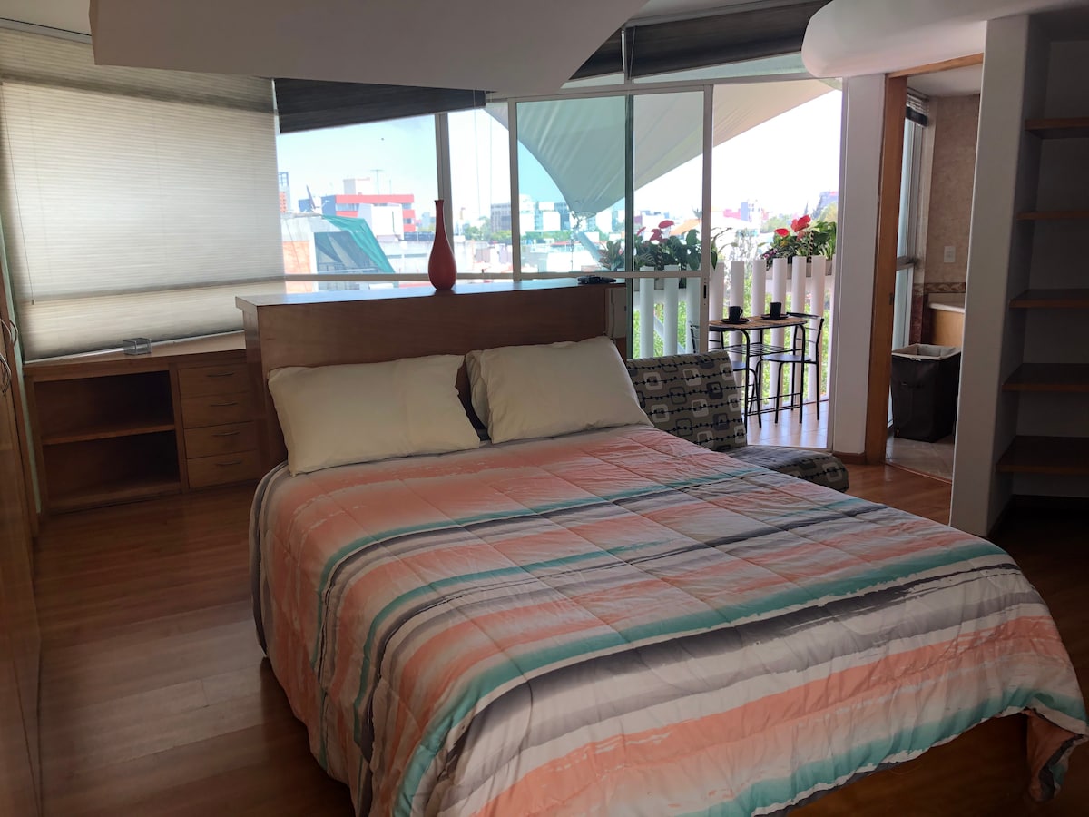 A spacious bedroom features a large bed with a striped blanket and natural light streaming in through floor-to-ceiling windows. There is a comfortable seating area with a single chair and a wooden cabinet, enhancing the room's functionality.