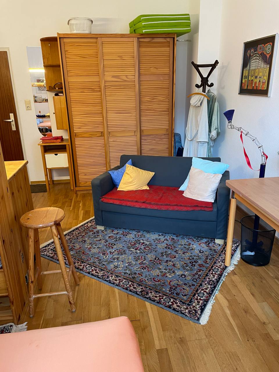 A cozy seating area features a positioned sofa adorned with colorful cushions. A decorative rug lies beneath, complementing the warm wooden flooring. Natural light highlights a simple wooden table nearby, and a wardrobe stands against the wall, providing additional storage.