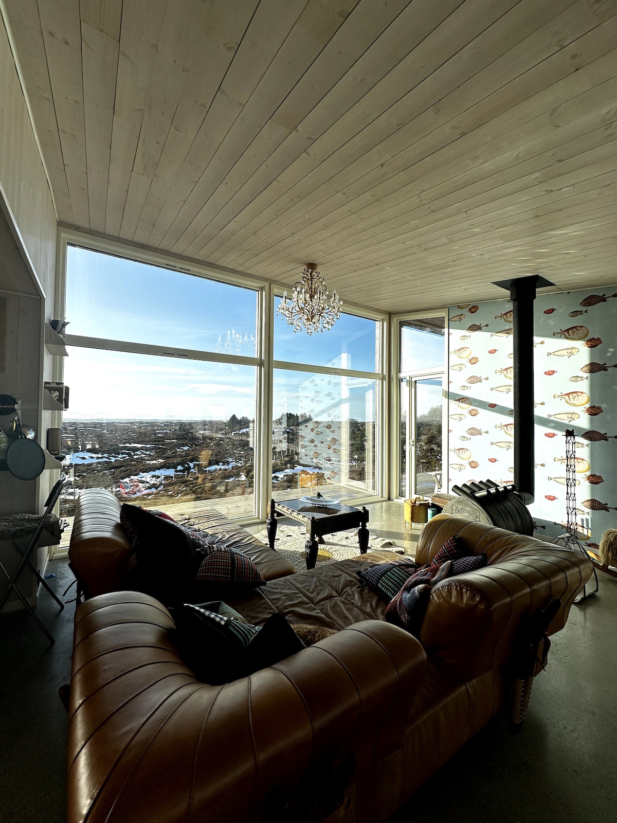 A bright living area features large windows that provide expansive views of the surrounding landscape. A chandelier hangs from the wooden ceiling, and a cozy leather sofa is arranged near a decorative wall. Light pours in, enhancing the inviting atmosphere.