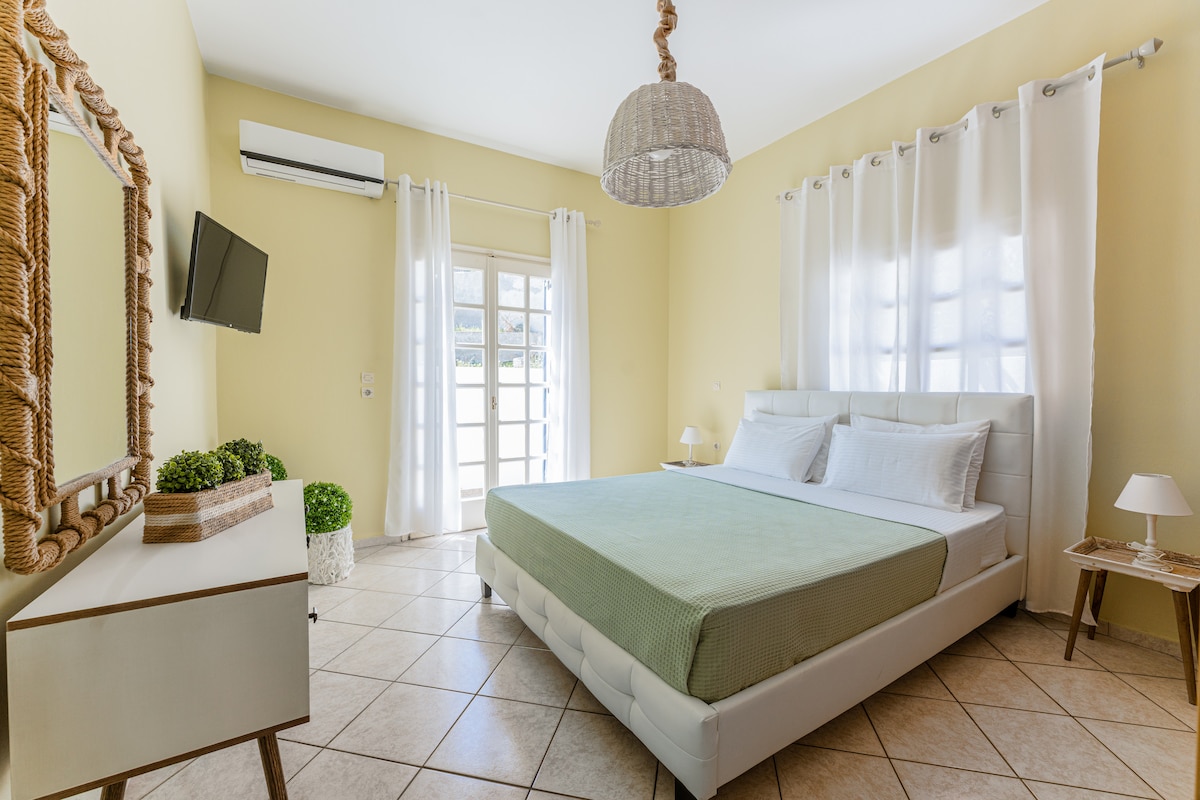 A modern bedroom is highlighted by a spacious bed with crisp white linens. Natural light filters through sheer curtains, illuminating a minimalist dresser and a small bedside table. A flat-screen TV is mounted on the wall, and an air conditioning unit is visible.