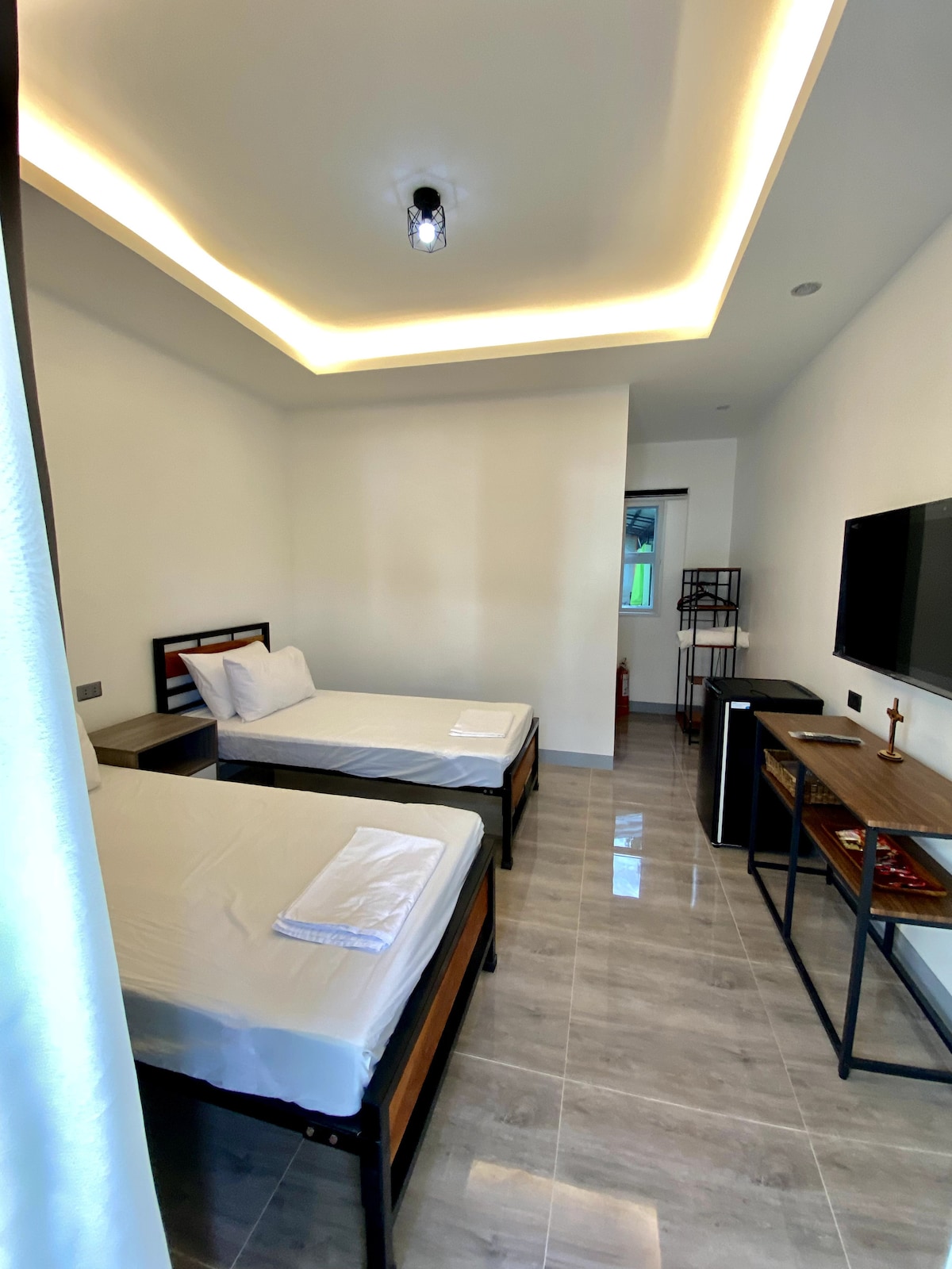 A bright and airy room features two single beds with crisp white linens. A wall-mounted smart TV is positioned opposite the beds, alongside a mini fridge and a wooden desk. Natural light enters through a window, enhancing the serene setting.