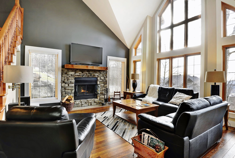 A spacious living area features large windows, allowing natural light to fill the room. A stone fireplace serves as a focal point, surrounded by comfortable leather seating. A wooden coffee table is positioned centrally, complemented by a patterned area rug and side tables with lamps.