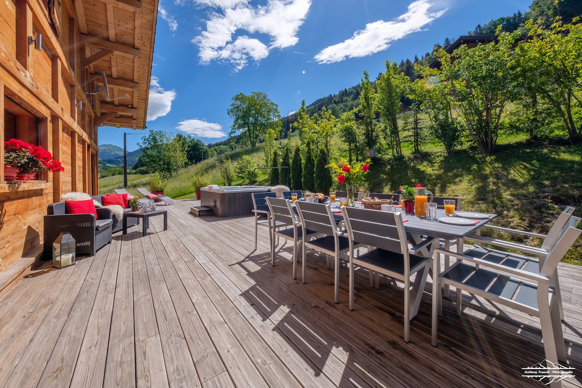 A spacious terrace is featured with a large dining table surrounded by multiple chairs, complemented by a seating area. A hot tub is visible, and sun loungers are arranged on the wooden decking. Lush greenery and a clear blue sky enhance the tranquil outdoor setting.