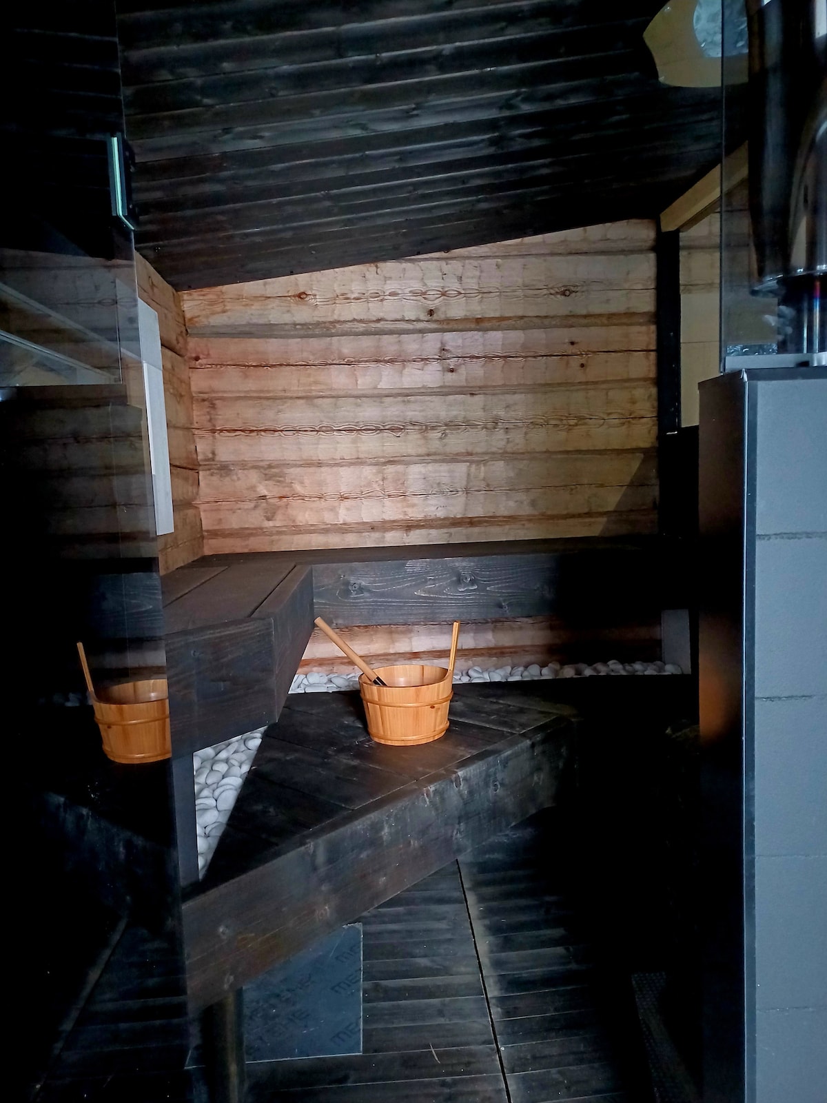 A serene sauna space features dark wooden walls and flooring, complemented by light-colored stones. A curved wooden bench is positioned against the wall, with a wooden bucket and ladle set nearby. The design promotes a calming ambiance, ideal for relaxation.