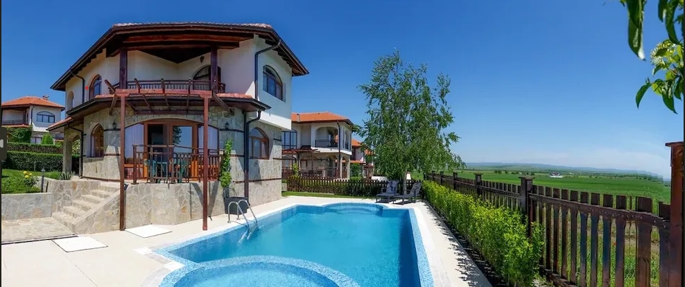 The outdoor area features a private pool surrounded by well-maintained landscaping. A spacious terrace overlooks the pool, complemented by comfortable seating and a water slide. The two-story villa showcases a light-colored facade and includes multiple balconies with wooden railings, enhancing the scenic views.