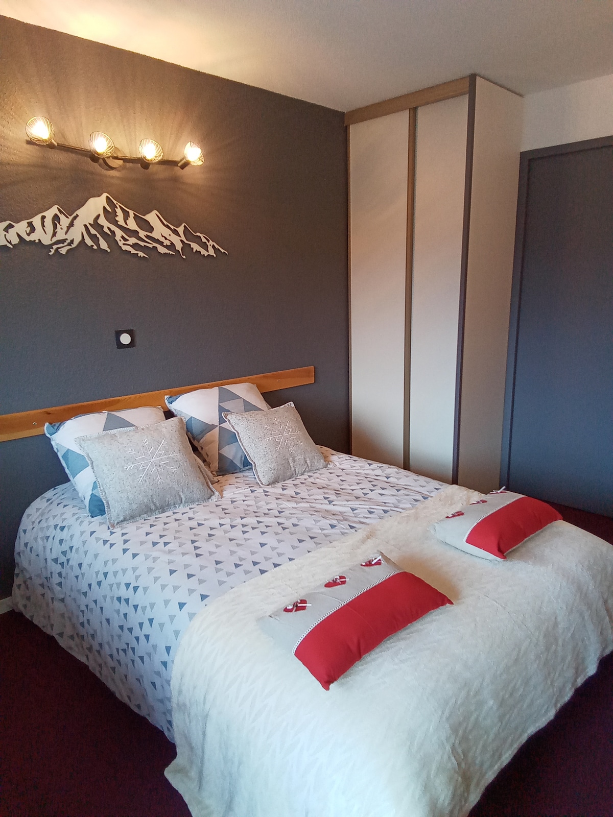 A sleeping area is furnished with a spacious bed featuring a patterned duvet and soft pillows. The walls are painted in a calming grey, and decorative lighting complements the room's ambiance. A large wardrobe provides additional storage space.
