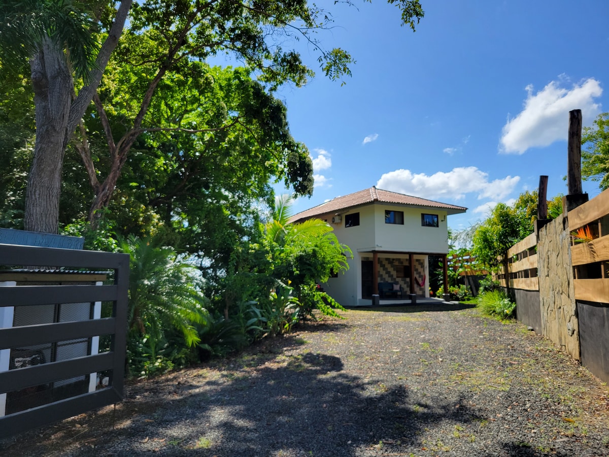The entrance to the property is lined with lush greenery and a gravel pathway leading to a two-story home. The modern structure features large windows and a welcoming design amidst a backdrop of blue skies and scattered clouds.