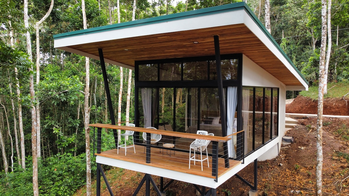 A modern bungalow is elevated on stilts, surrounded by lush greenery. The structure features large glass windows and sliding doors that offer expansive views of the forest. A wooden terrace is furnished with outdoor chairs, blending seamlessly into the natural environment.