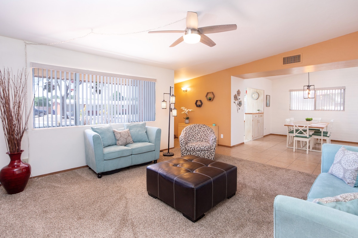 A spacious living area features light walls, a ceiling fan, and large windows allowing natural light. Comfortable seating includes a turquoise sofa and a patterned chair. A dark ottoman centers the space, while a dining area is visible in the background.