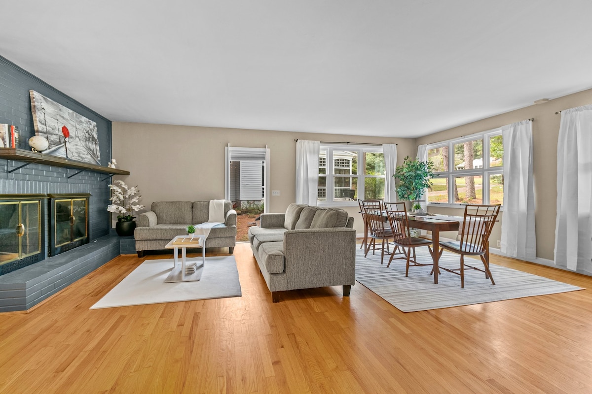 A spacious living area features a cozy seating arrangement with soft gray sofas and a light wooden coffee table. Natural light filters through large windows adorned with sheer white curtains, creating an inviting atmosphere. A dining table with wooden chairs is positioned nearby, on a light area rug.