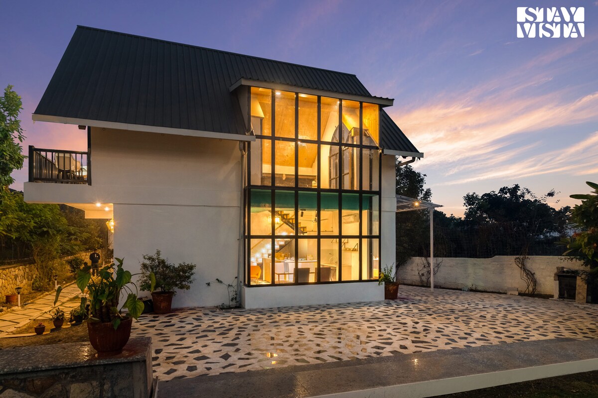 A modern duplex cottage is showcased under a twilight sky, with large, illuminated windows that reflect the warm light from inside. The exterior features a stone-paved patio surrounded by potted plants, enhancing the connection between indoor and outdoor spaces.