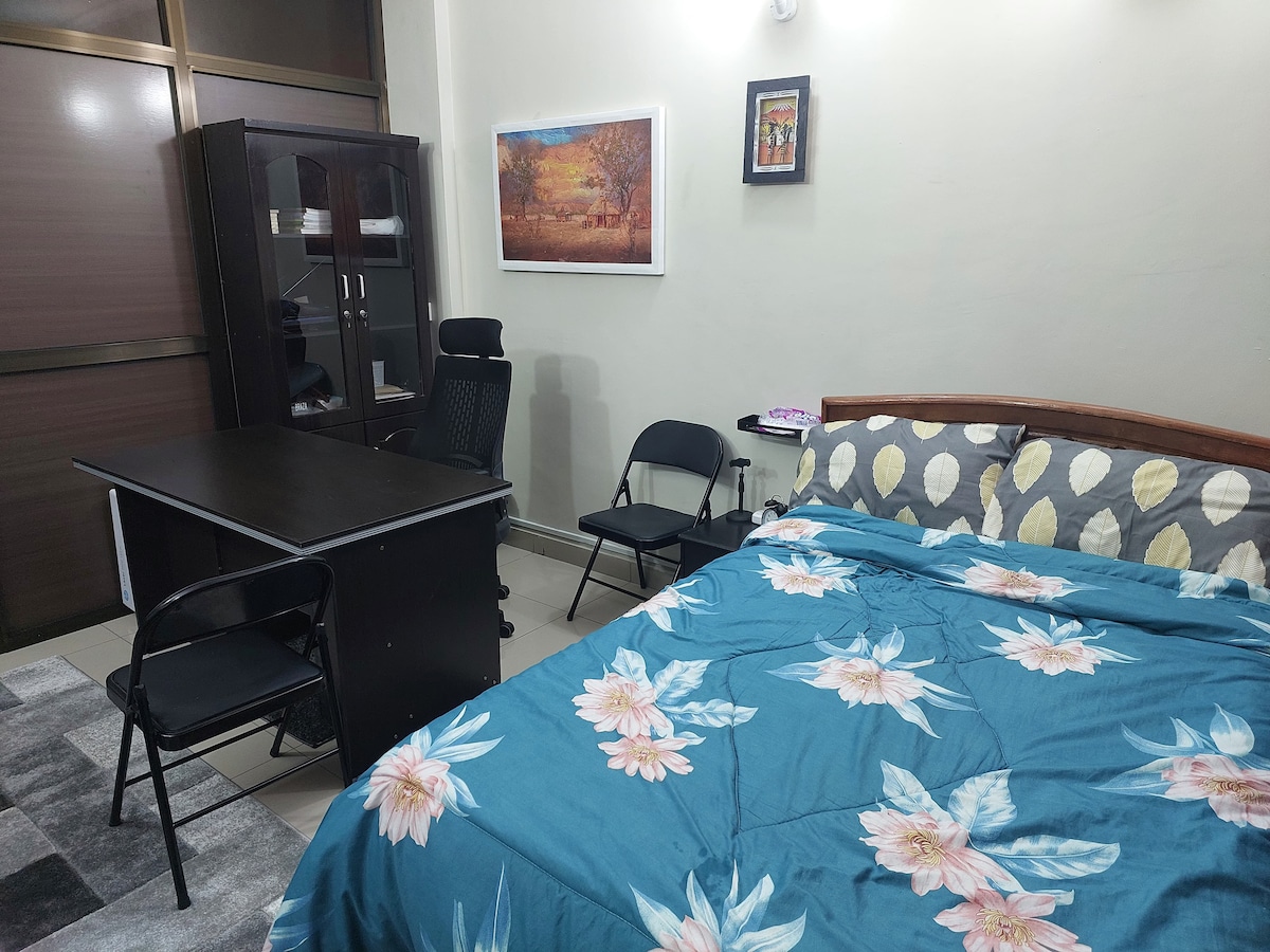 The room features a spacious bed adorned with floral bedding, complemented by a desk and an office chair. A black shelf unit stands to the side, displaying a framed artwork. Soft lighting allows the calming colors to enhance the serene atmosphere.