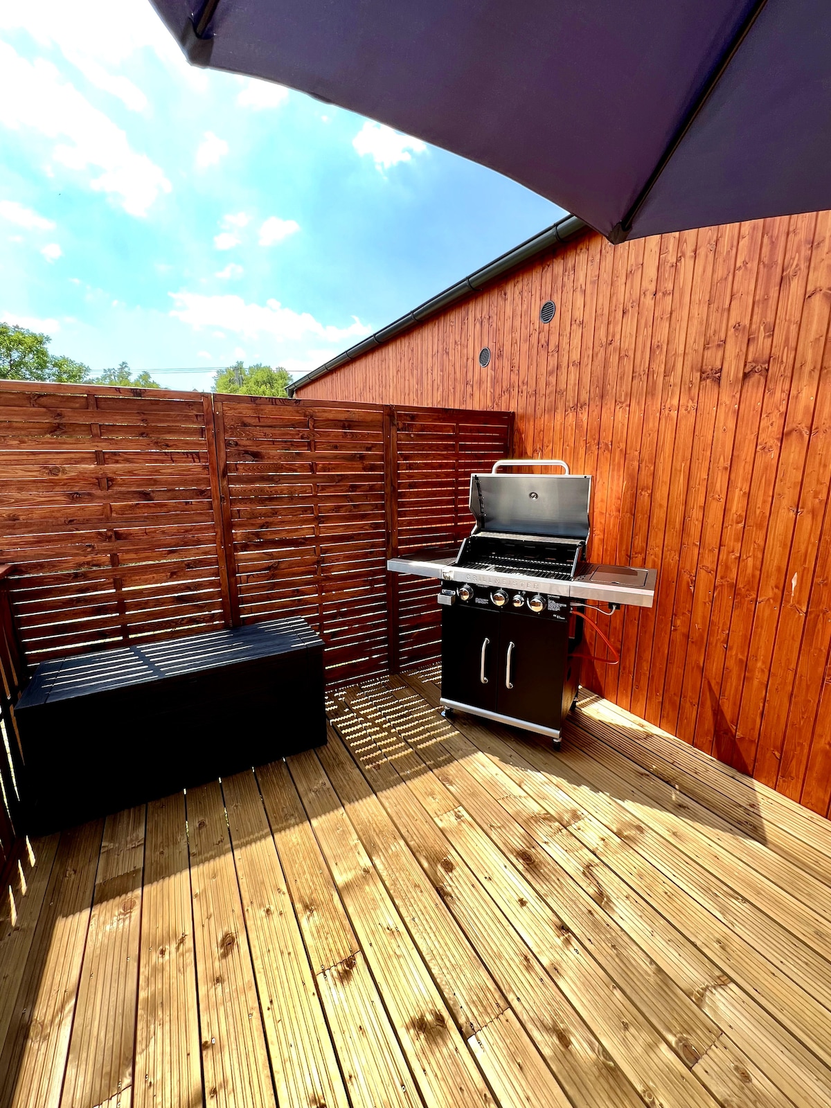 A private terrace is featured with wooden decking and a shaded area. A black grill with three burners is positioned on the side, next to a storage bench. The surrounding wooden slats provide privacy and a cozy atmosphere.