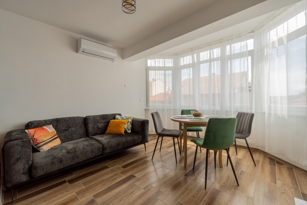 A well-lit living area features a dark gray sofa accompanied by a green and a wooden dining chair. A round dining table sits at the center, and large windows bathe the space in natural light, offering a view of the surrounding rooftops.