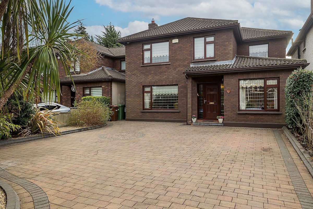 A brick house with a spacious driveway is shown, featuring large windows and a welcoming front door. The area is surrounded by greenery, including palm trees and shrubs. A light-colored vehicle is parked on one side, enhancing the sense of space.