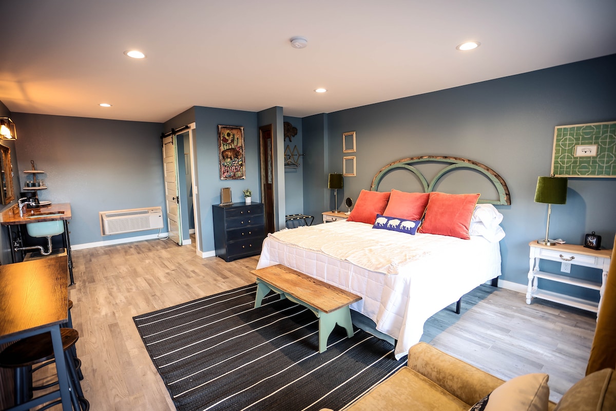A stylish room features a spacious bed with decorative pillows and a wooden bench at its foot. A workspace with a metal barstool is visible, along with a dresser and a cozy seating area. Light-colored flooring complements the blue walls and modern decor.
