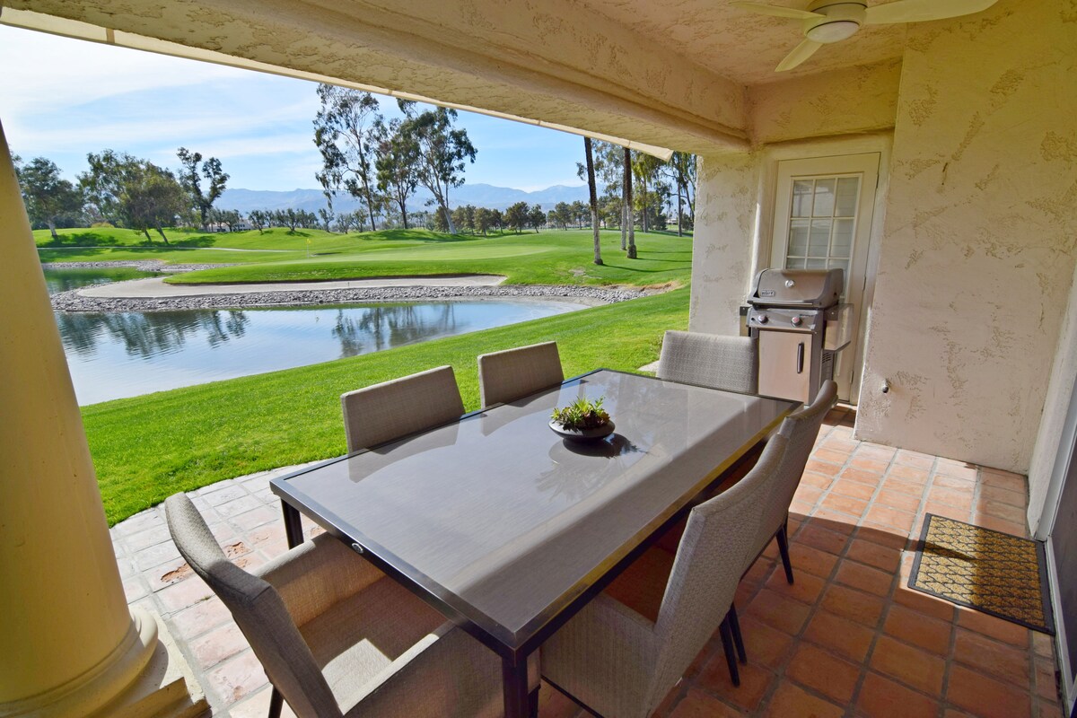 A private patio area features a dining table with six chairs, shaded by the overhanging structure. An outdoor grill is positioned nearby, with a scenic view of the landscaped yard and the lake beyond. Large sliding glass doors provide access to the interior.