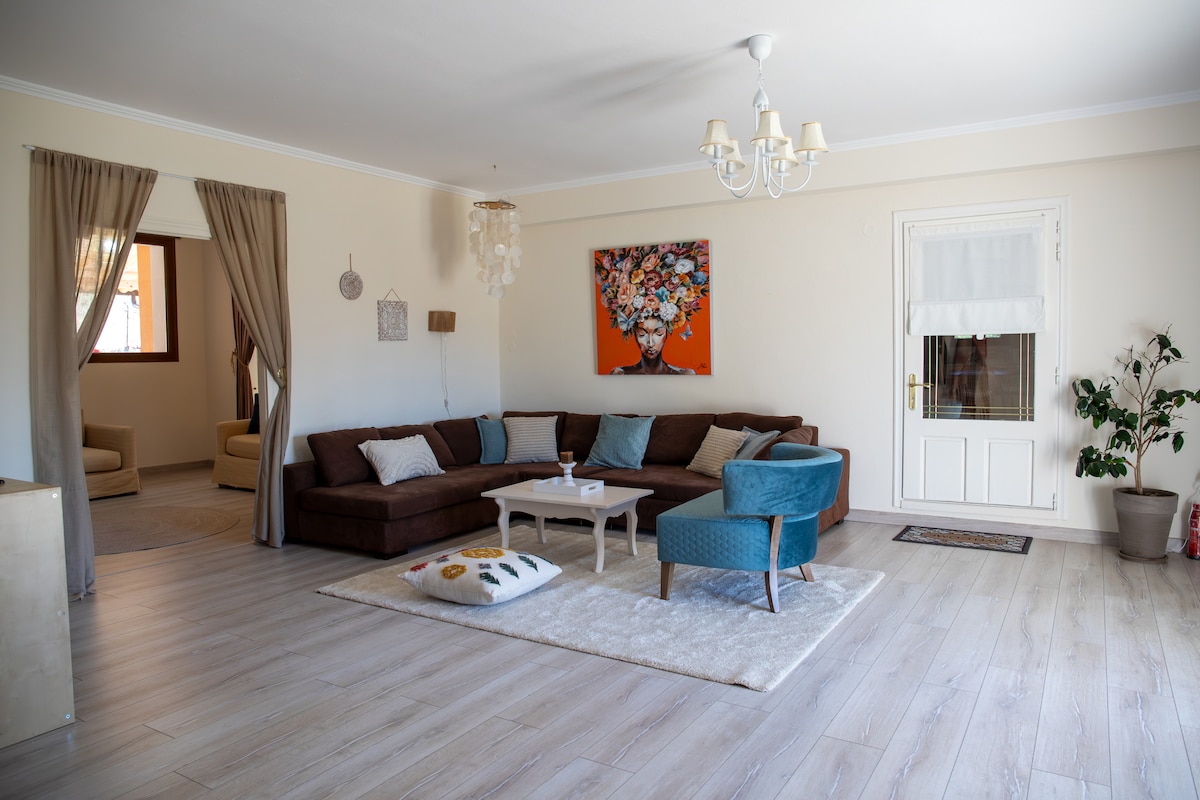 A spacious living area features a large brown sectional sofa complemented by colorful cushions. A light-toned coffee table sits in the center on a soft area rug. Natural light enters through windows, illuminating the neutral-toned walls and enhancing the cozy atmosphere.