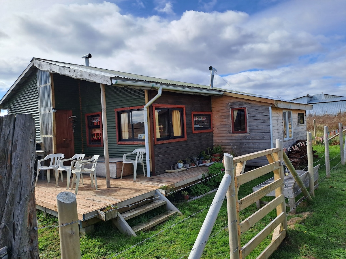 A rustic cabin is presented with a wooden deck and a small seating area, featuring several white chairs. The exterior combines dark green and wood paneling, while large windows offer glimpses of the surrounding nature.