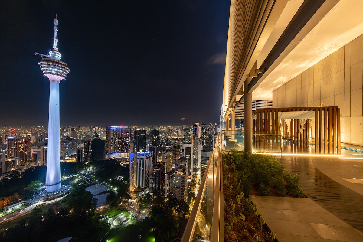 A panoramic night view highlights the vibrant skyline of Kuala Lumpur, featuring the iconic communications tower illuminated against the dark sky. The balcony is adorned with lush greenery, softly lit pathways, and modern architecture, providing an inviting space for relaxation.