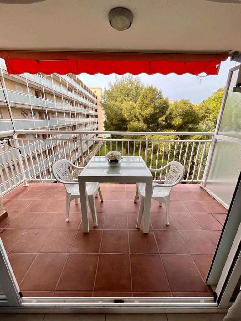 A small outdoor balcony is displayed, featuring a white table surrounded by two matching chairs. A red awning provides shade, while green trees are visible in the background. The tiled floor enhances the space, offering a cozy setting for relaxation.