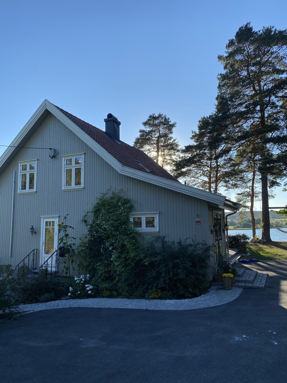 The exterior of a light-colored house is shown, framed by tall trees and open space. A paved path leads to the entrance, while a glimpse of water is visible in the background. Large windows reflect the sunlight, enhancing the home’s inviting presence.