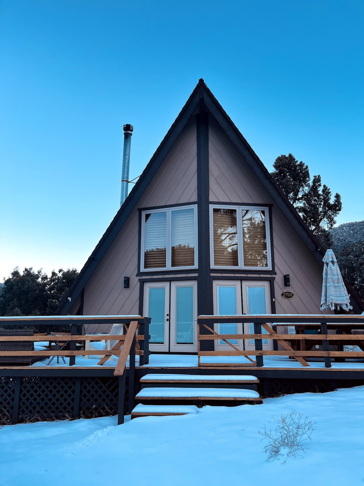 The A-frame cabin features a peaked roof and large windows, allowing natural light to flood the space. A wooden deck with a railing extends toward the entrance, surrounded by snow. A patio umbrella is visible, adding a touch of outdoor comfort.